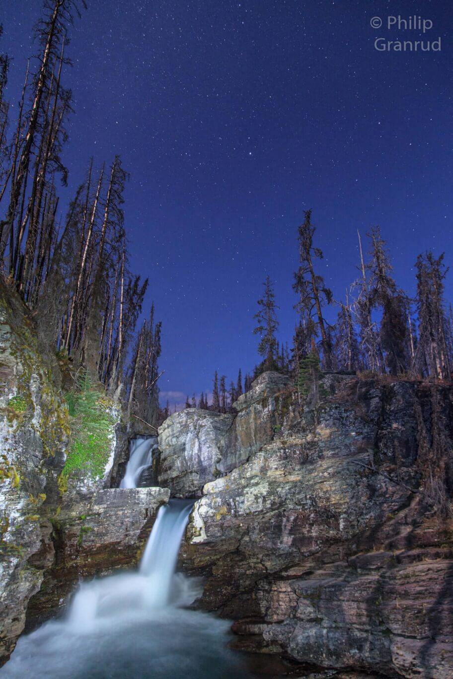 Night sky with stars in Glacier National Park with a waterfall and cliffs in foreground